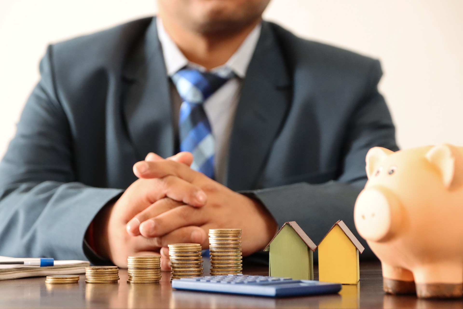 Close-up image of unrecognisable business man wearing smart, blue suit with white, button down collared shirt and blue checked tie sat behind desk, stacked coins, calculator, piggy bank, cardboard houses, real estate and finance concept