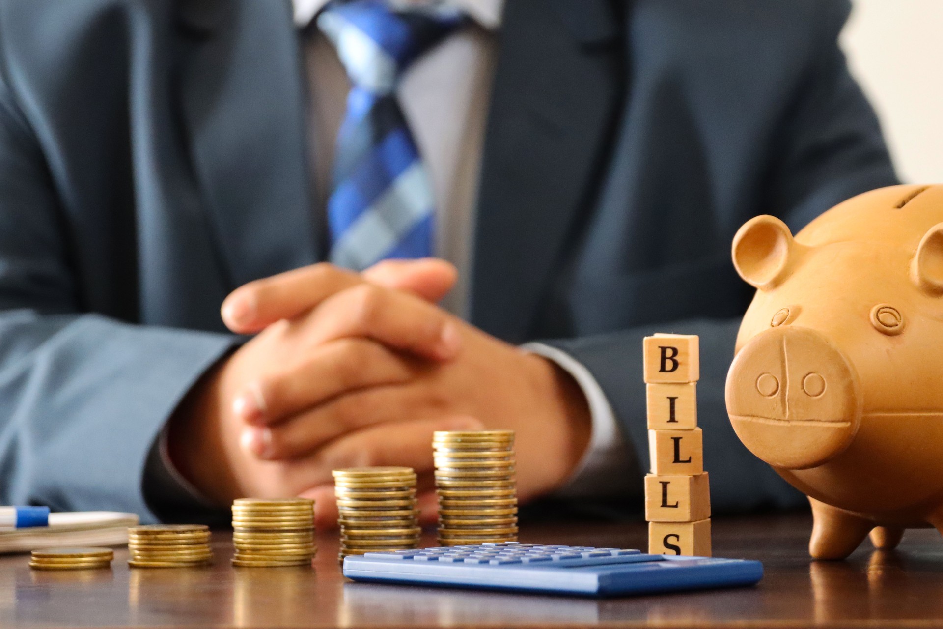 Close-up image of unrecognisable business man wearing smart, blue suit with white, button down collared shirt and blue checked tie sat behind desk, stacked coins, calculator, piggy bank, wooden cubes spelling bills, banking and finance concept